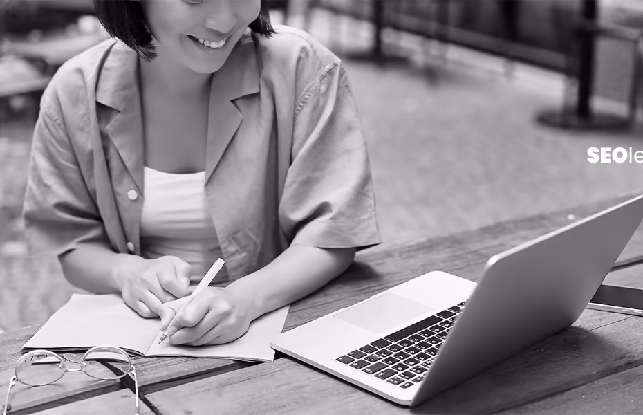 A woman writing in a notebook beside a laptop at an outdoor café, representing content marketing and blogging work for digital content creation.